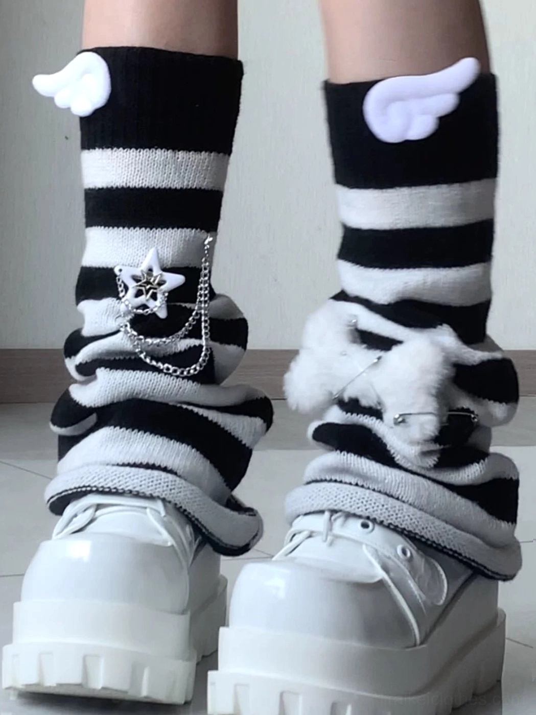 Full close-up of calf and feet wearing bold black and white striped leg warmers with angel wings layered star chain and fluffy pompom with all-white platform shoes