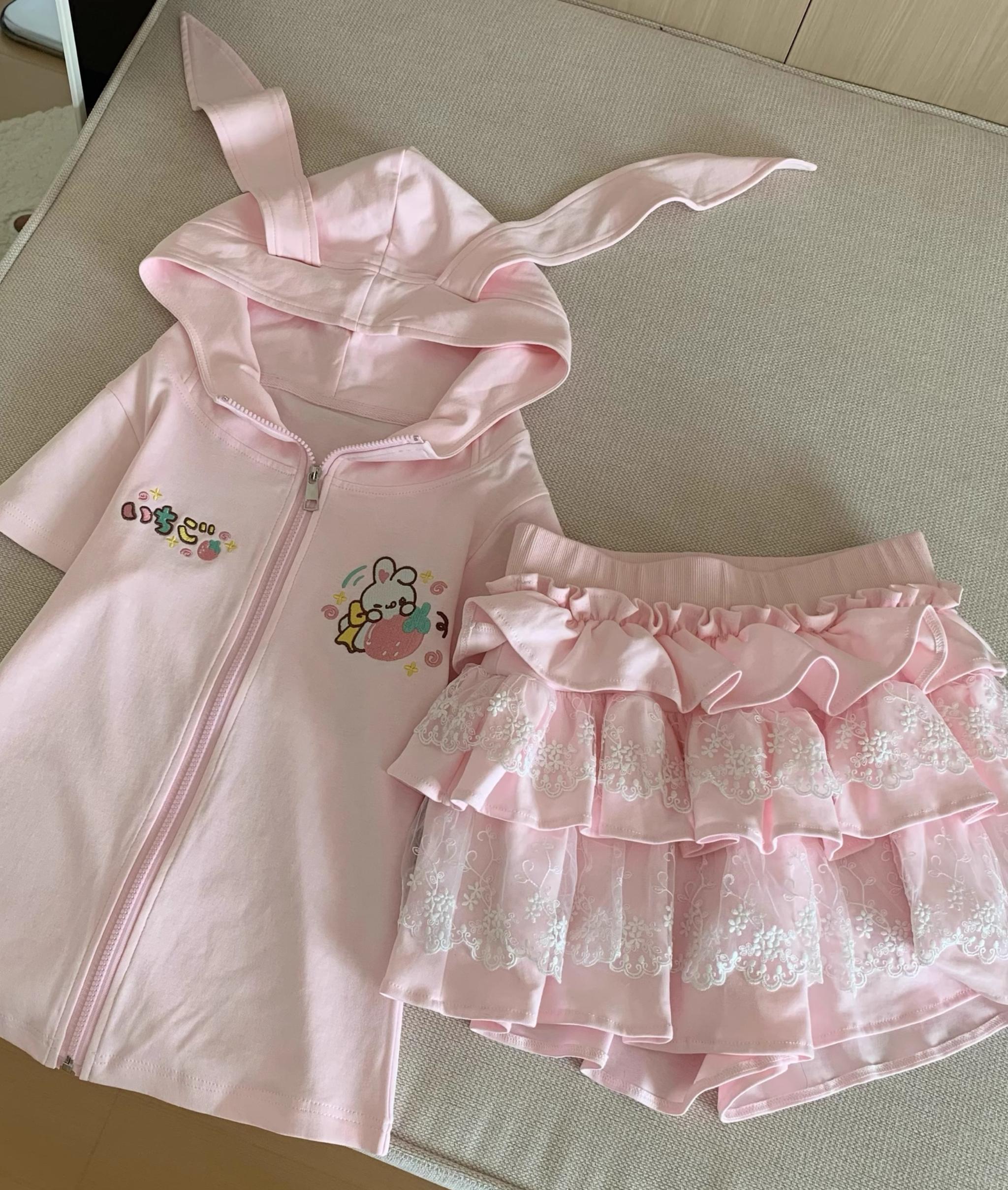 Light pink bunny-ear zip-up hoodie with three-tier lace ruffle cake skirt separated two-piece set flat lay on beige cushion