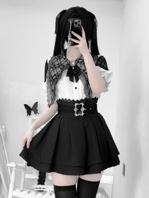 Jirai Kei lace capelet blouse and high-waist pleated skirt set worn by model with twin tails, featuring white ruffle blouse with black lace capelet and mini skirt with belt, styled with black thigh-high stockings in home setting