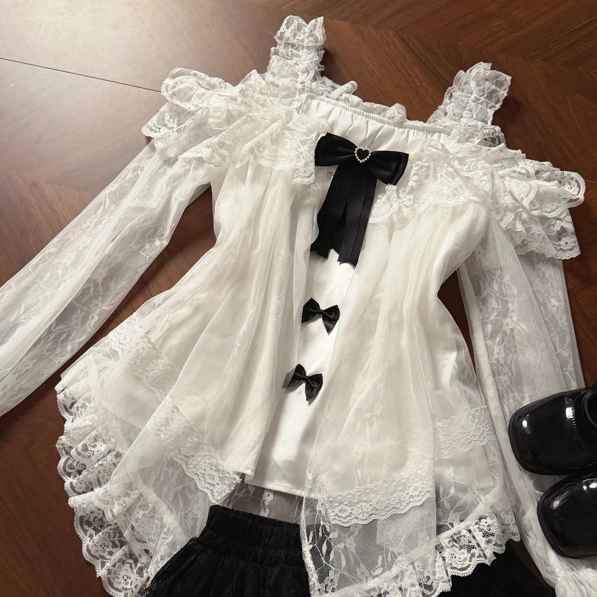 Flat lay top-down view of off-shoulder black-and-white lace blouse displaying multiple removable satin bows, delicate ruffled cuffs, and intricate lace hem trim on wooden surface