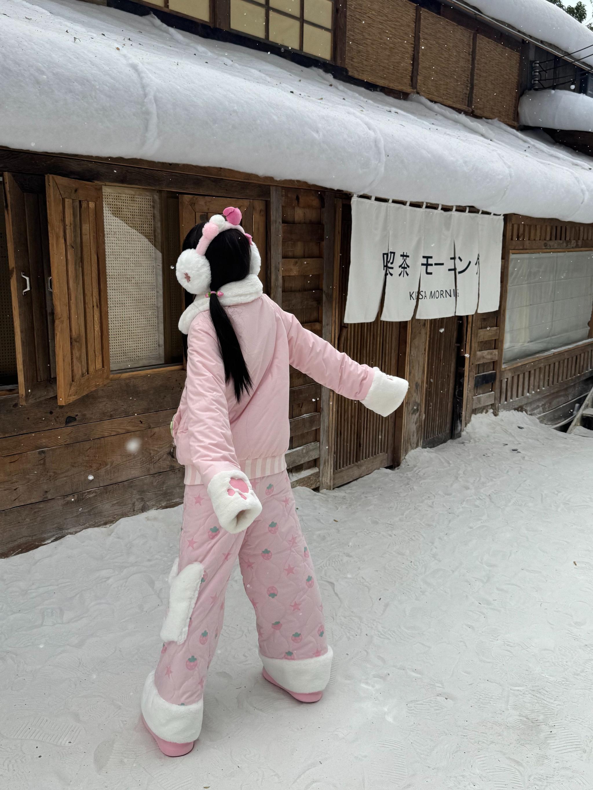 Side back full-body view of pink Jirai Kei quilted winter outfit featuring fuzzy earmuffs matching fuzzy-trimmed jacket all-over fruit prints and white ankle cuffs on snowy wooden Japanese street