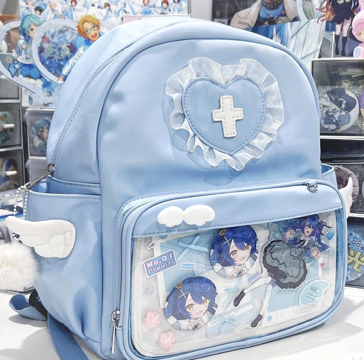 Light blue Jirai Kei itabag backpack front-right oblique angle showing grid-patterned transparent display pocket with anime goods, angel wing details and heart cross badge