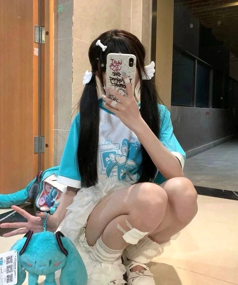 Low-angle squat mirror selfie featuring cyan and white raglan sleeve cat ear hooded anime tee on long black twin-hair otaku model, paired with white ruffle skirt and plush toys