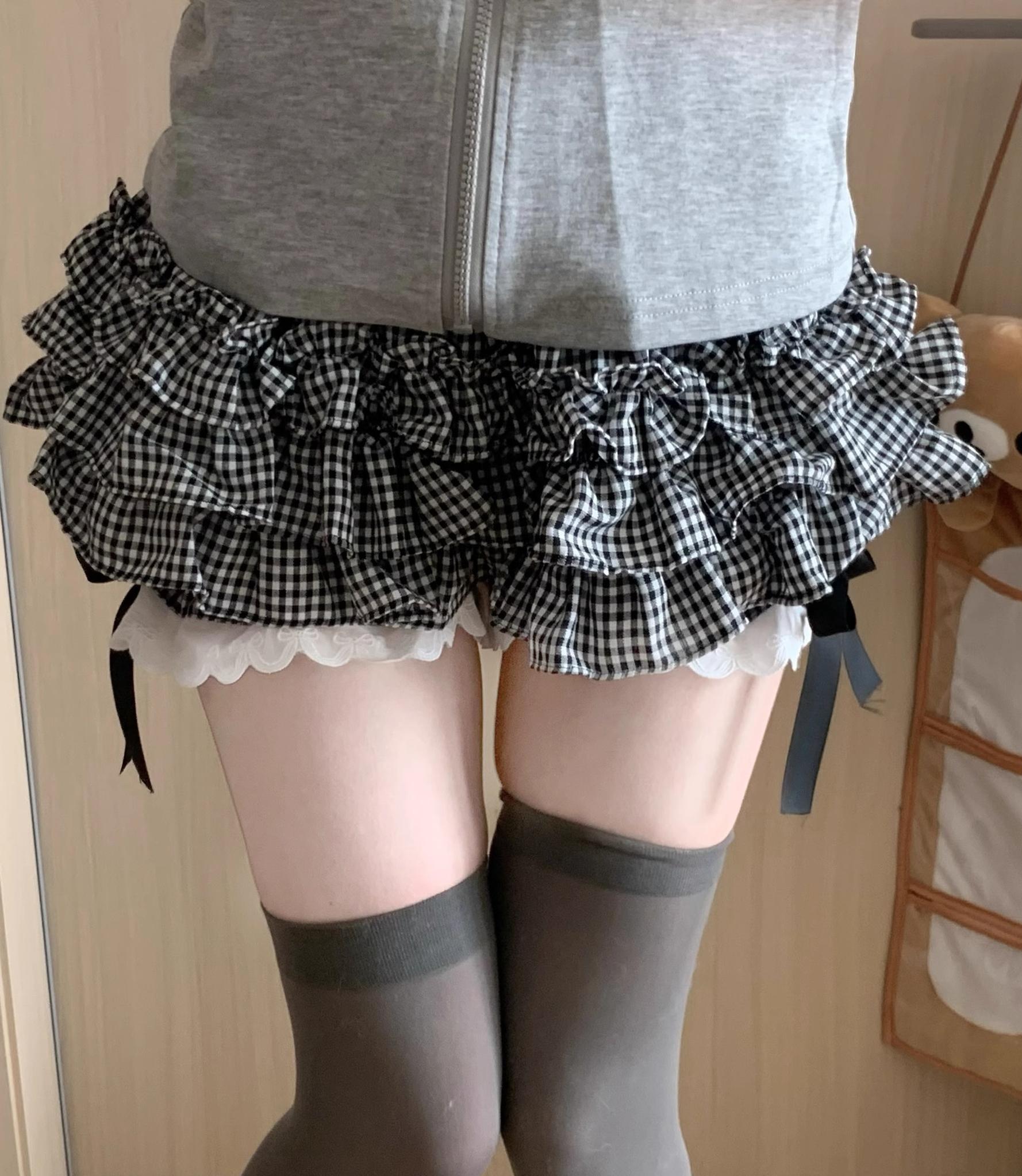 Black and white gingham plaid Jirai Kei mini skirt close-up detail shot featuring ruffled layers white scalloped lace trim built-in shorts and side bow accents styled with grey thigh-high socks