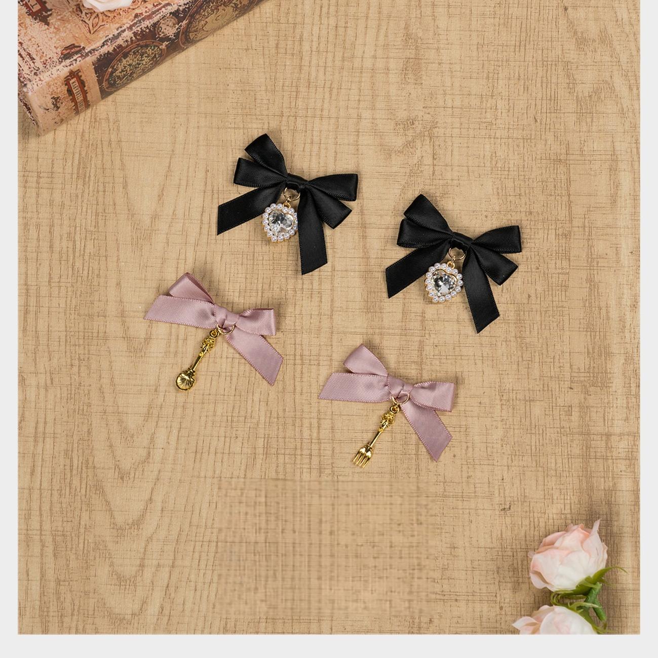 Black and light pink Jirai Kei bow hair clips with rhinestone and bell charm pendants flatlay arrangement on wooden table with artificial flowers