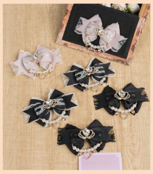 Delicate lace Jirai Kei bow clips set in dusty pink and black with rhinestone crown, pearl chain and heart charm details displayed on wooden table flat lay