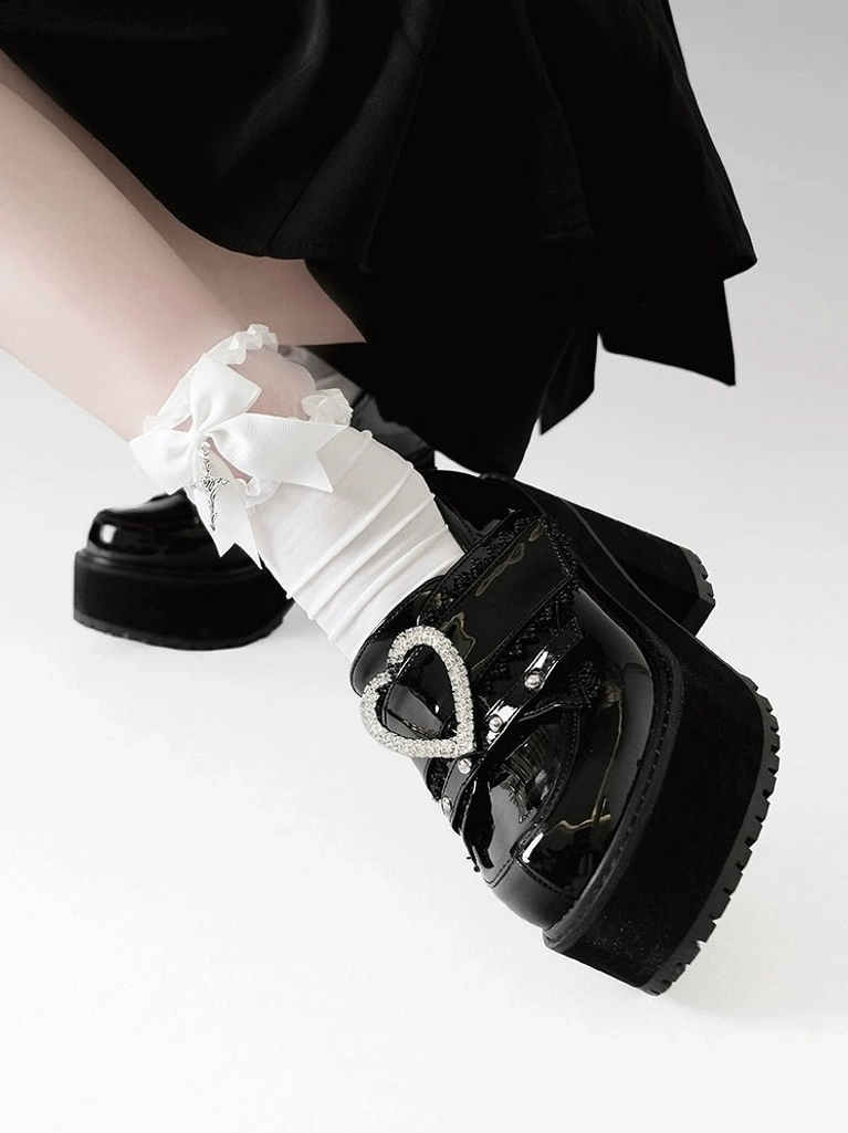 Low side close-up single foot view of large rhinestone heart buckle platform loafers with thick block heel, patent leather finish, partial jirai kei skirt and frilled bow socks visible