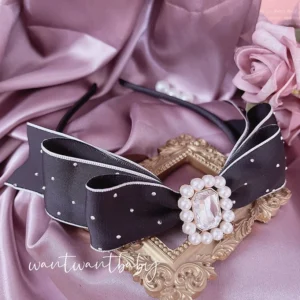 Black dotted double-bow Jirai Kei KC headband with white pearl and clear rhinestone center, flat lay on pink satin cloth with decorative elements