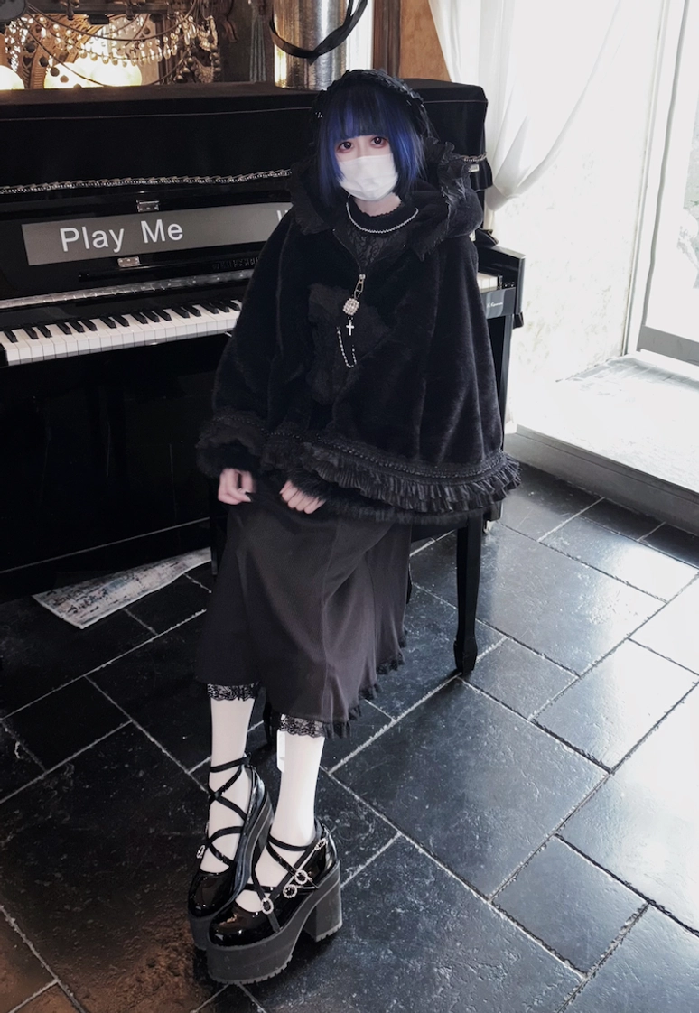 Left three-quarter front view of black fleece cape coat with cat ear hood at piano cafe, styled with cross strap platform shoes and satin midi skirt