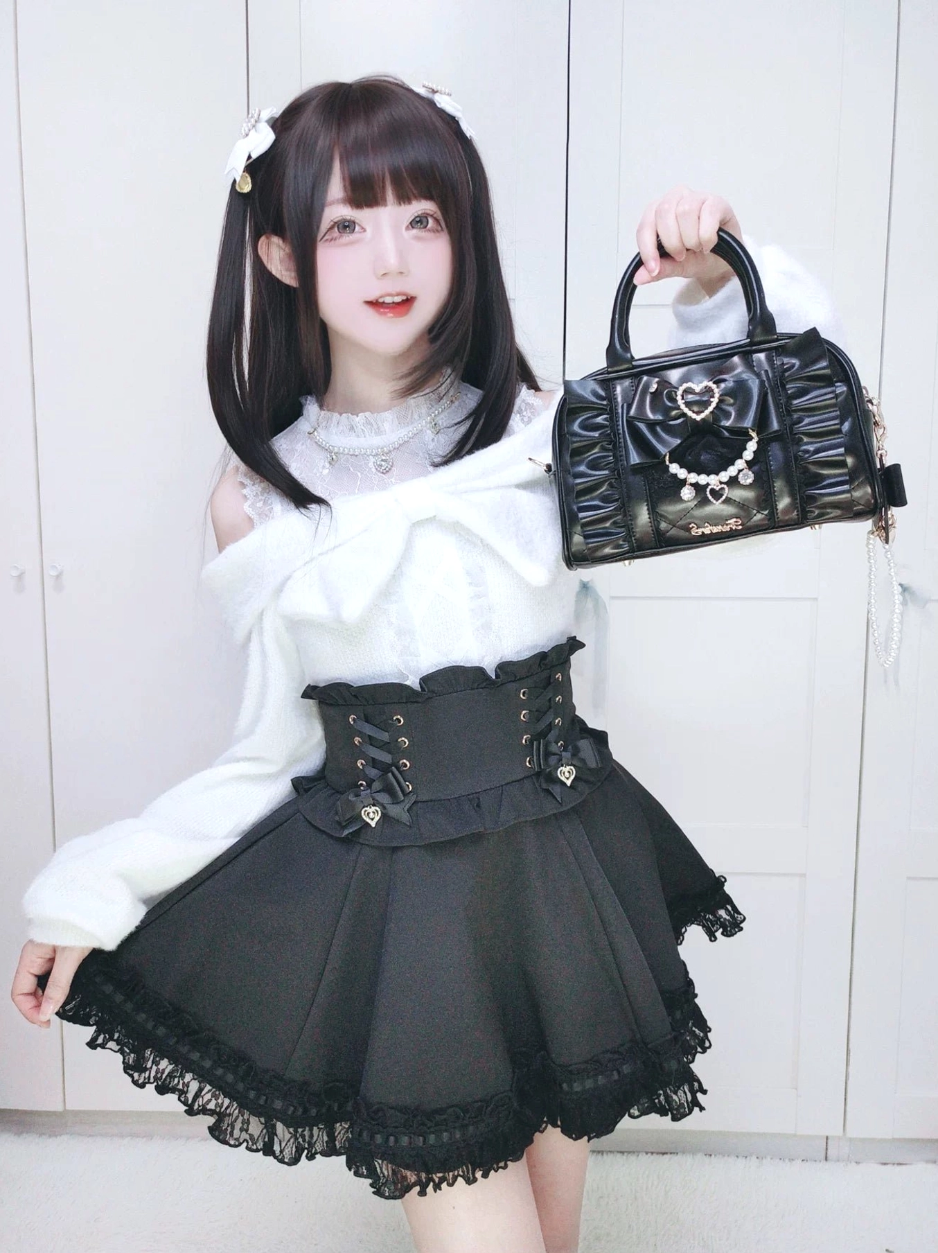 Model wearing sweet Lolita outfit with black Jirai Kei handbag, twin ponytails hairstyle, indoors white door backdrop styling shot