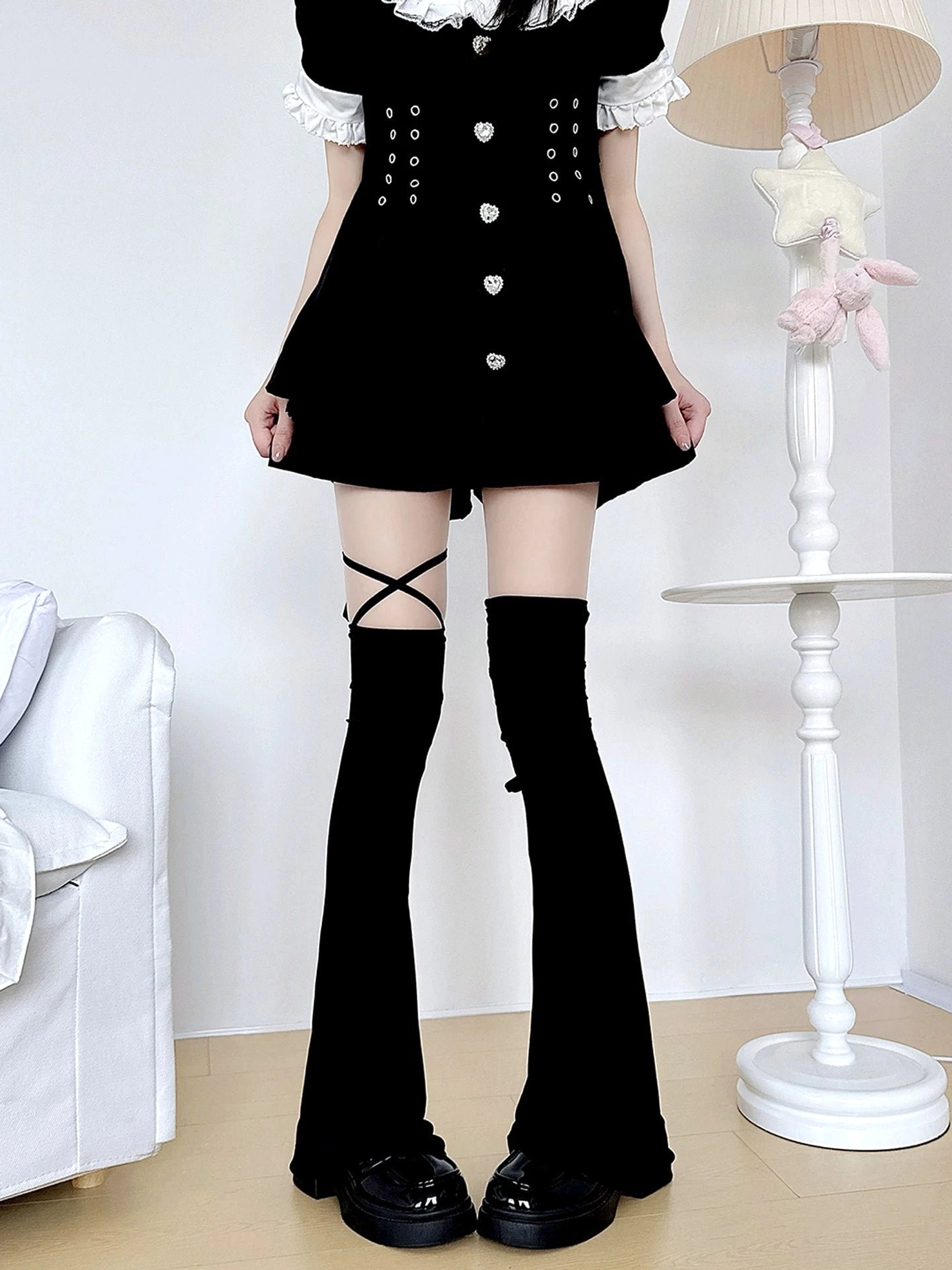 Black jirai kei over-the-knee leg warmers with adjustable cross straps worn with black lolita mini dress, featuring flared hem and thigh straps in indoor home setting