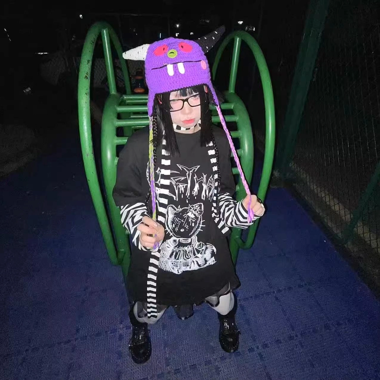 Full-body front view of black jirai kei striped false two-piece long sleeve punk tee with black-white striped sleeves and cartoon cat print, worn at indoor amusement space