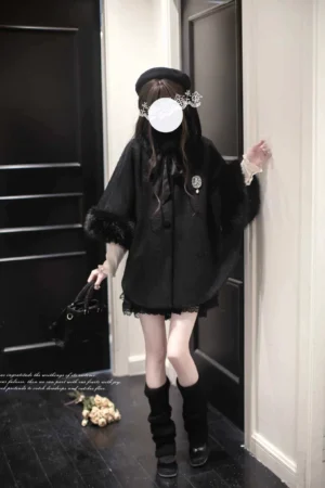 Black jirai kei winter cloak with faux fur trim worn by female model in indoor entryway, featuring decorative brooch and elegant hand-tied bow collar