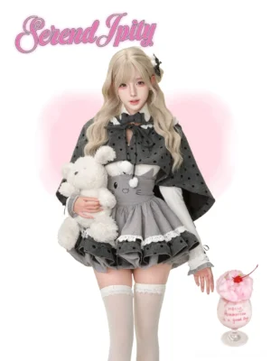 Jirai Kei dark angel blonde model wearing complete cat-themed Lolita outfit with polka dot cape, white knit top with Juliet sleeves, and gray cat-face embroidered suspender skirt with ruffle layers against pink heart backdrop, full-body front view