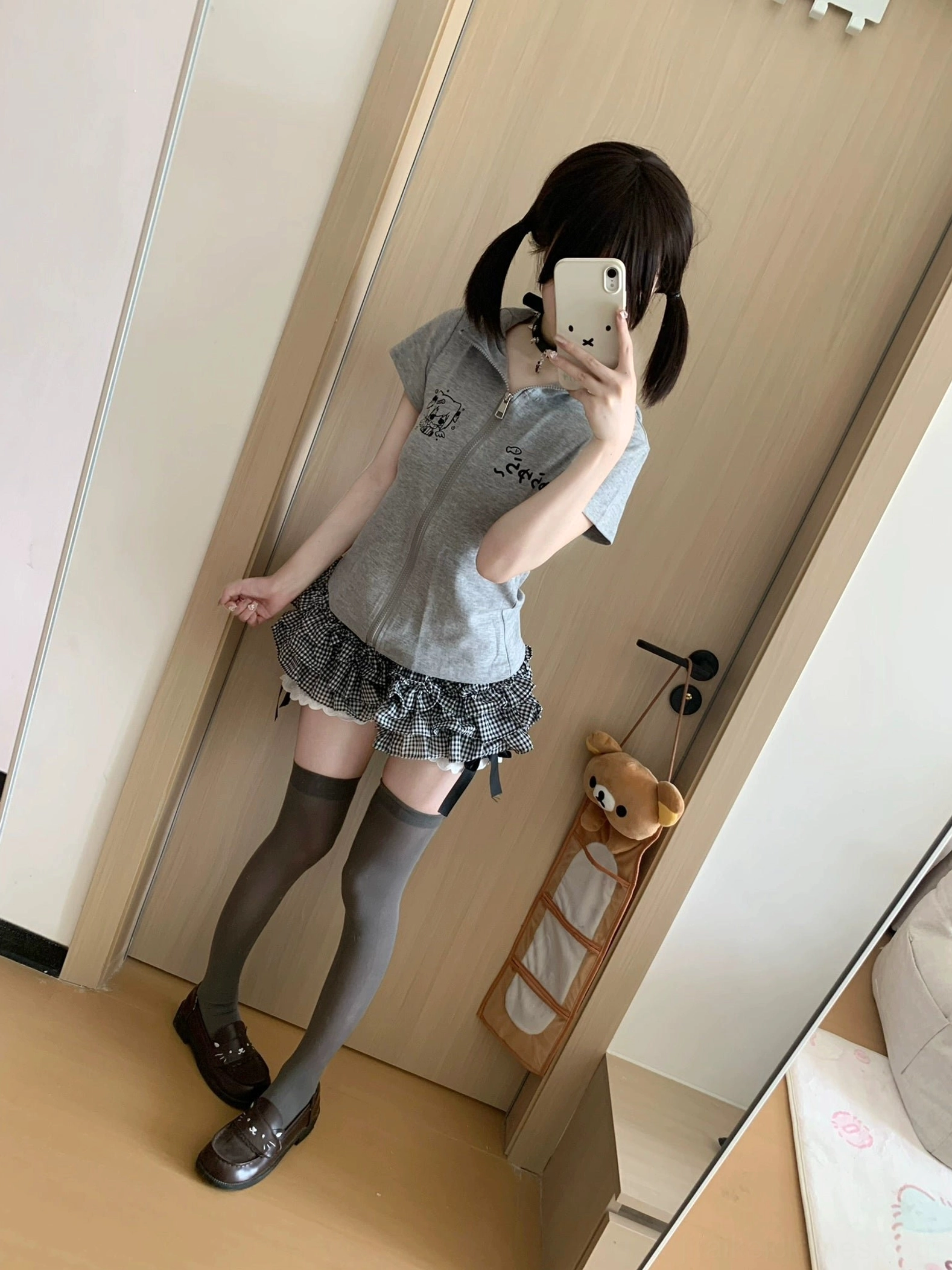 Full-body mirror selfie modeling complete Jirai Kei outfit featuring black white plaid mini skirt with built-in shorts grey zip-up hoodie with cartoon prints matching loafers thigh-high socks and twin-tail hairstyle bedroom aesthetic