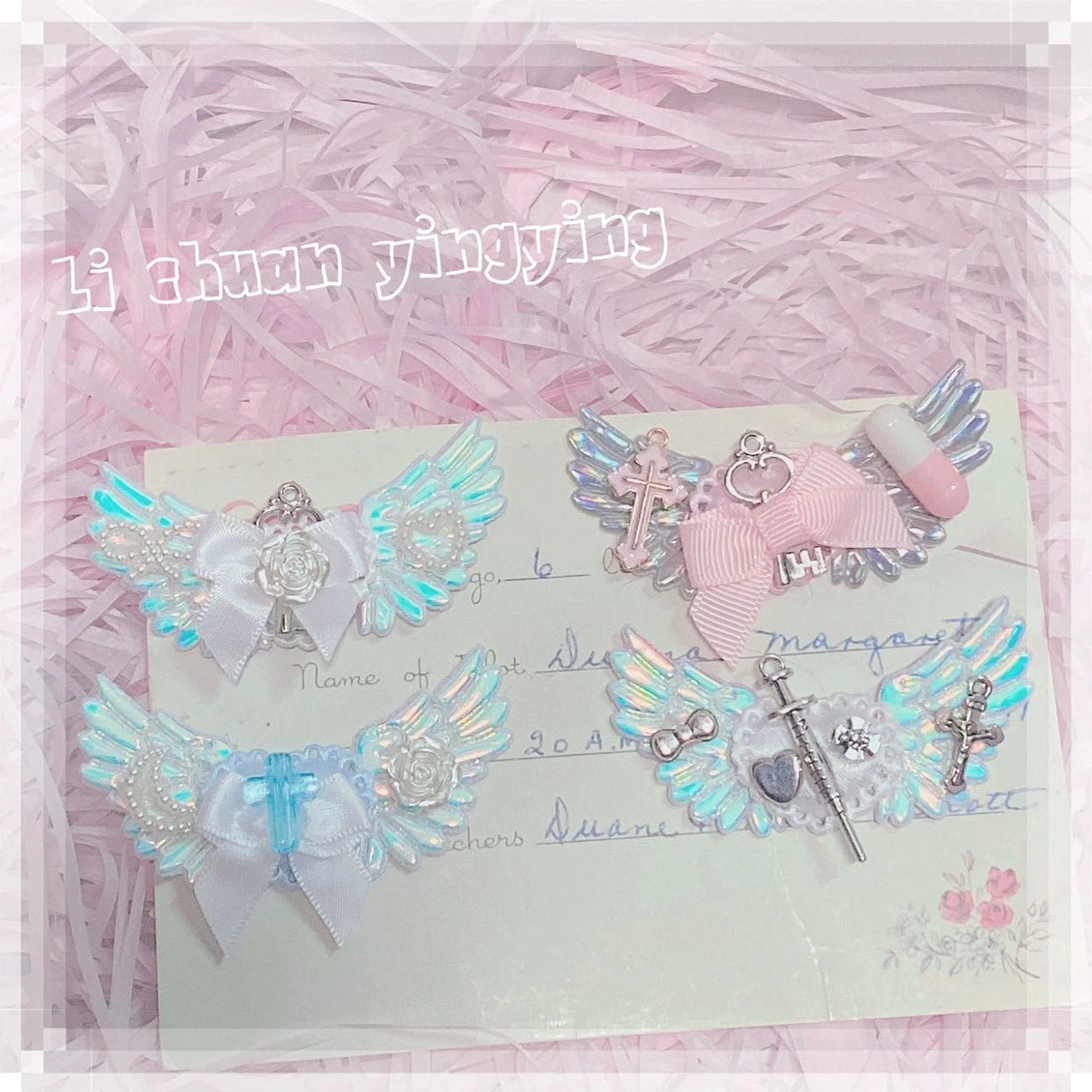 Assorted jirai kei hair clips arrangement on pink shredded paper with display card showing blue white tones iridescent wings cross and pill decorations