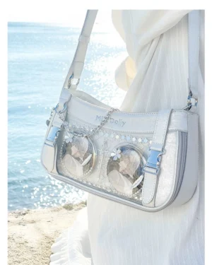 Side close-up of light silver-blue Jirai Kei ita shoulder bag worn on model at seaside beach, showing dual clear badge pockets, branded shoulder strap, and shiny holographic PU leather texture