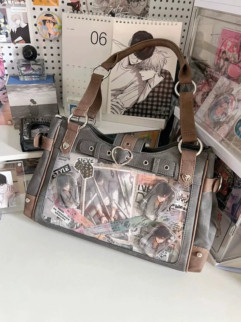 Gun-grey and denim blue two-tone Jirai Kei ita shoulder bag with transparent display panel filled with anime stickers and collectibles, shown on anime-themed desk backdrop