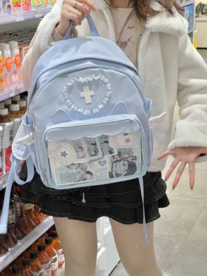 Blue Jirai Kei itabag backpack with angel wing decorations and heart lace cross badge worn by model in convenience store setting, showcasing transparent display window with anime merchandise
