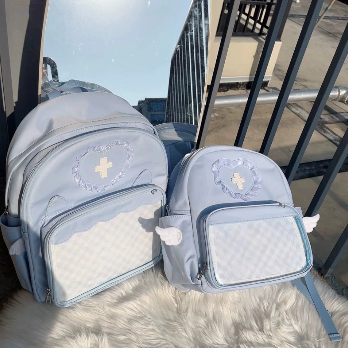 Two sizes of light blue Jirai Kei itabags displayed together on rooftop metal floor with fur mat, showing both S and L size transparent display pockets with grid lining and angel wing accents
