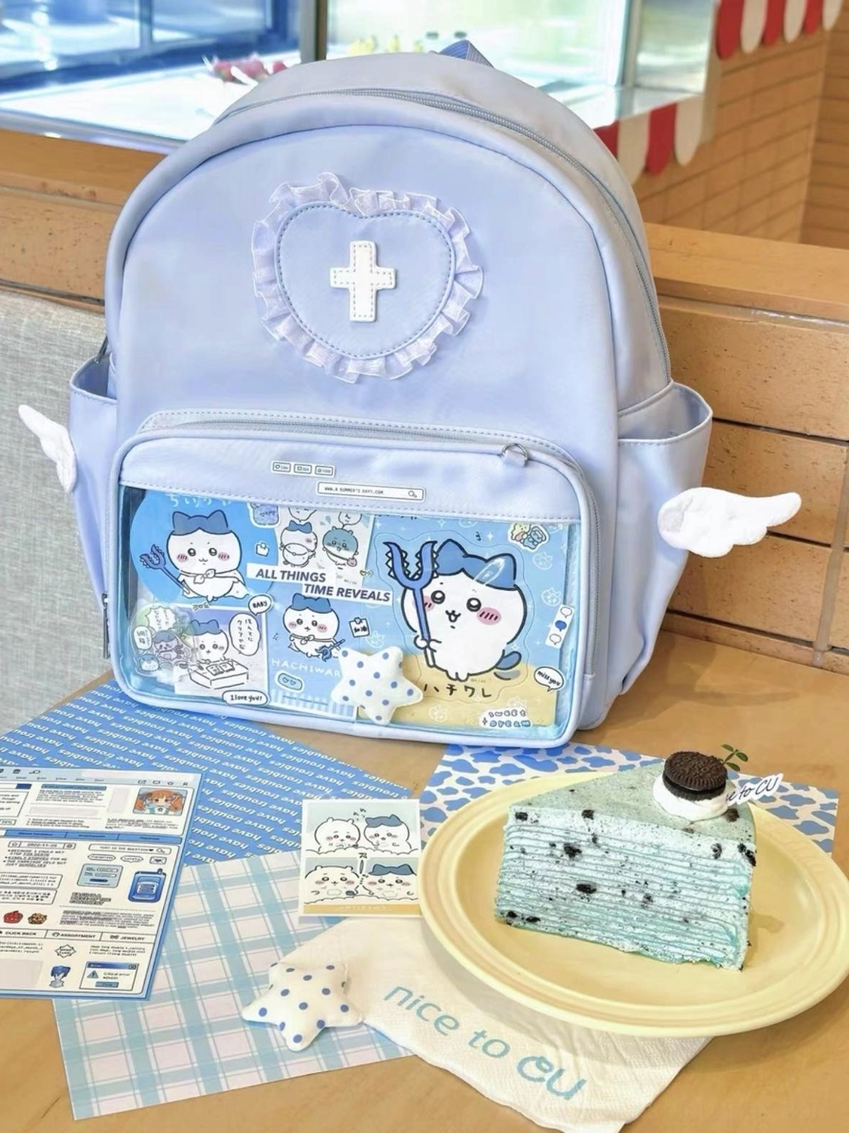 Top-down oblique view of blue itabag backpack at afternoon tea desktop scene, displaying transparent pocket with Chiikawa anime figures, heart lace cross emblem and angel wing side decorations