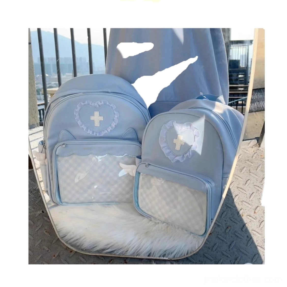 Two light blue Jirai Kei itabackpacks right oblique front angle view against rooftop metal railing with city skyline, showing heart lace cross decorations, grid clear display pockets and angel wing accents