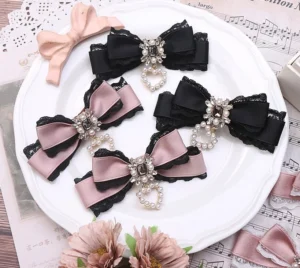 Double layer Jirai Kei lace bow hair clips in black and nude pink color options displayed on white ceramic plate with sheet music aesthetic background, showcasing rhinestone and pearl chain details