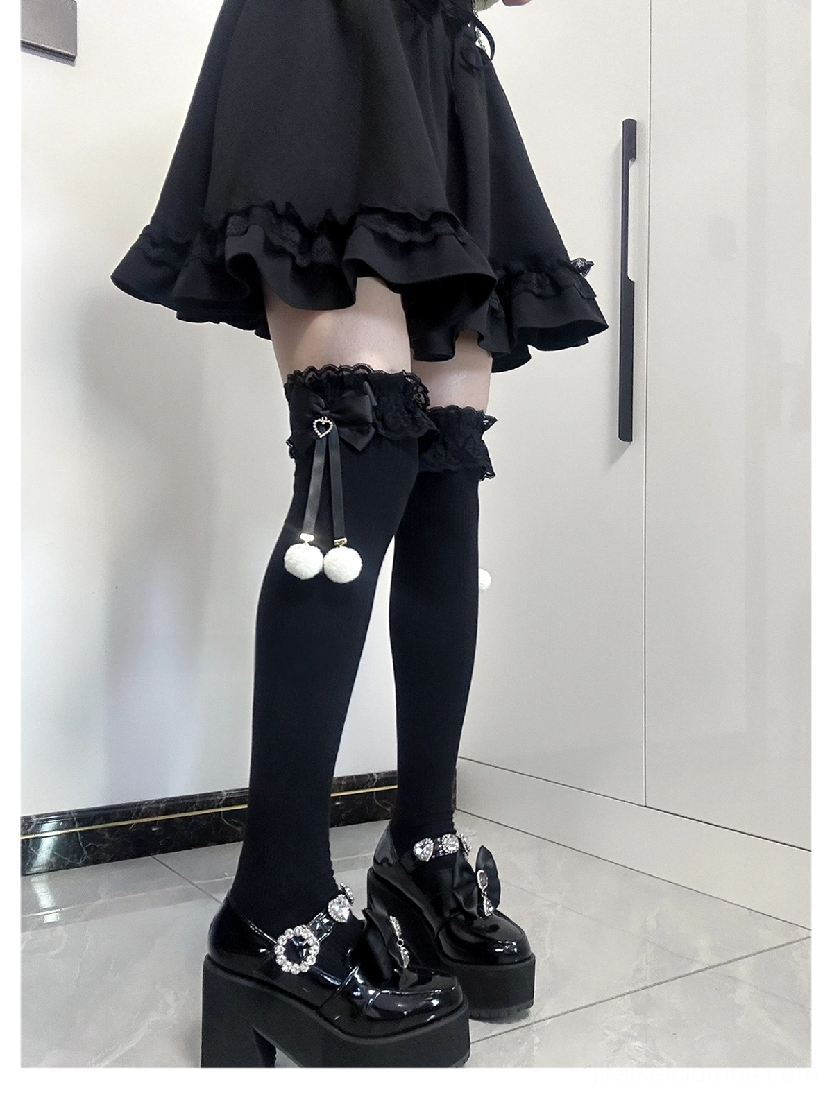 Jirai Kei thigh-high socks three-quarter side view all-black solid socks with lace cuffs black bow accents and white pom-pom pendants hanging details worn with black ruffled skirt