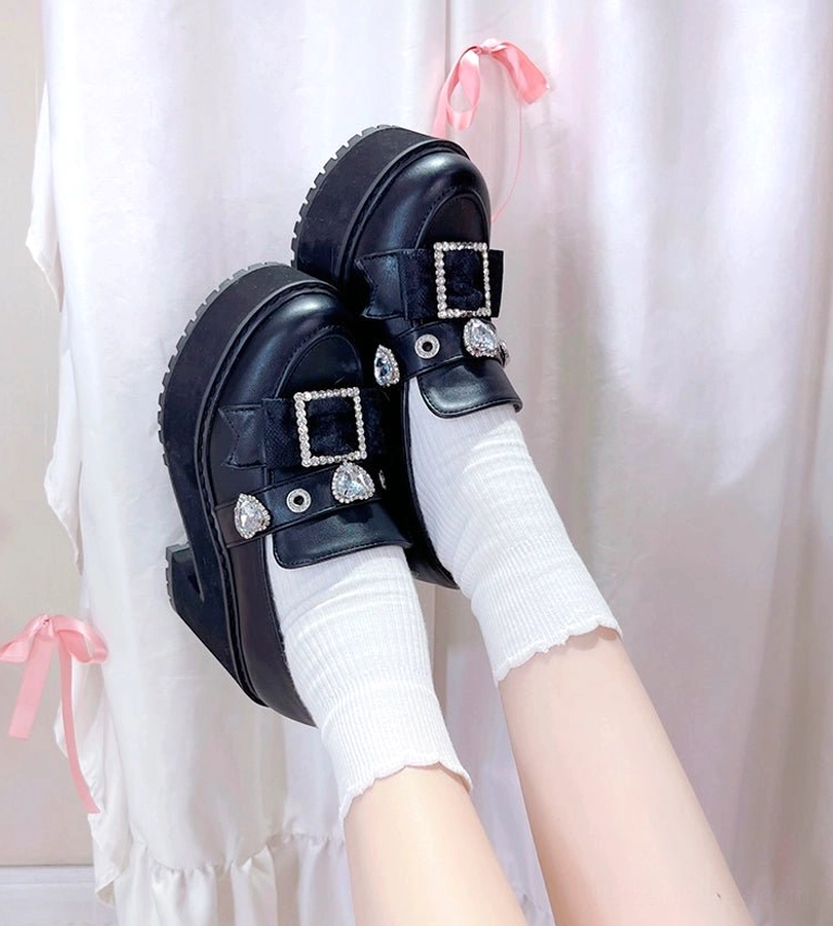 Angled close-up of black Jirai Kei loafers featuring heart-shaped rhinestone accents paired with white ribbed ankle socks against pink bow backdrop
