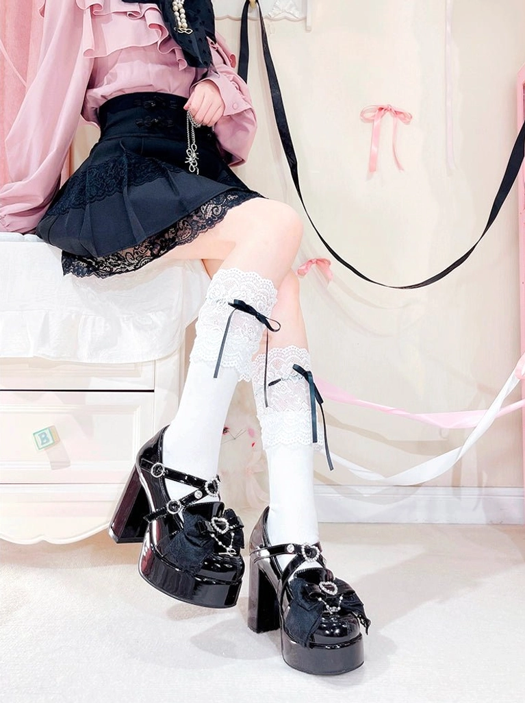 Low-angle side view of black Jirai Kei lolita platform heel shoes on seated model, showing thick platform sole, heart rhinestone ornament, and delicate bow vamp decoration