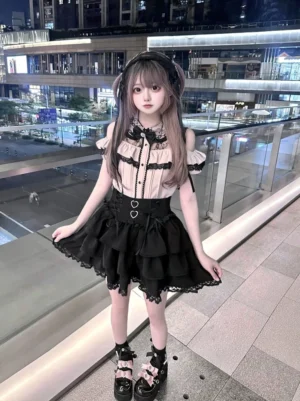 Jirai Kei off-shoulder blouse with tiered black lace skirt worn by young woman at night outdoor shopping mall terrace, featuring apricot-black color scheme and layered ruffled skirt with heart waist details