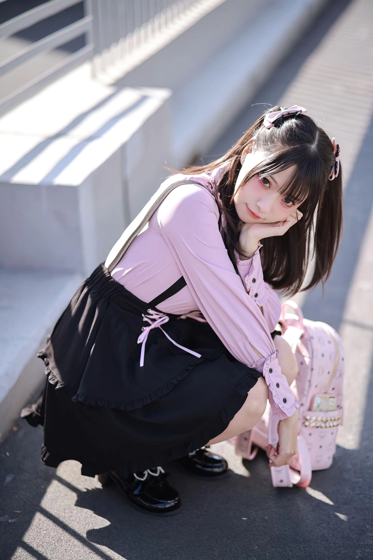 Outdoor casual Jirai Kei coordinate side-squatting view featuring twin-tailed girl, black suspender skirt with pink lace-up, leather flat loafers, outdoor stair setting
