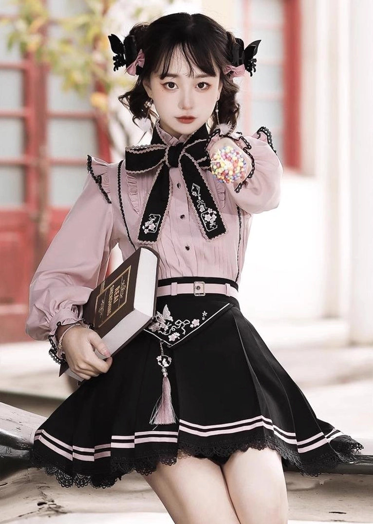 Full body low-angle shot of pink and black Jirai Kei two-piece set with model holding vintage book, showing lace-trimmed skirt hem and European street background