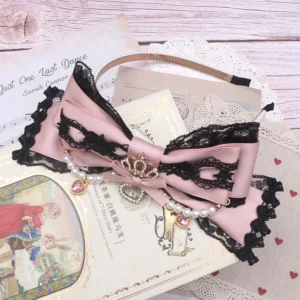 Pink-black Jirai Kei lace bow headband with crown charm and pearl heart rhinestone pendants, flat lay on vintage wooden desk with antique book and doily