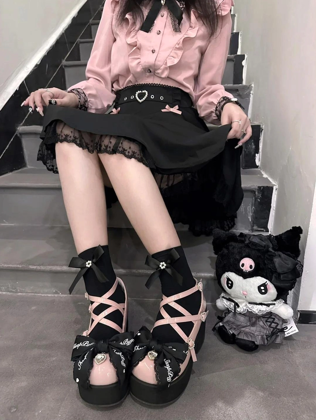 Frontal squatting view of pink and black platform Mary Jane shoes with heart-shaped belt buckle, ruffled lace skirt visible, coordinating bows, and Kuromi plush toy aesthetic styling