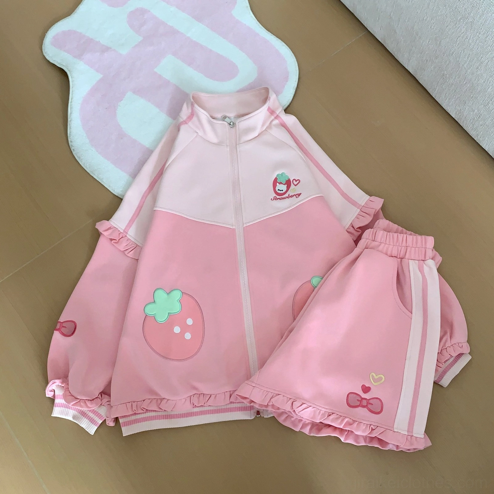 Top-down flat lay of pink sporty Jirai Kei two-piece set, showing side stripe shorts detail with heart embroidery and matching strawberry-themed coat with ruffled elements