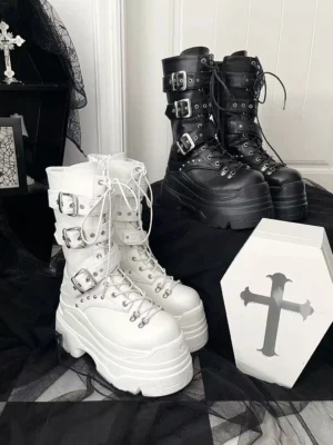 Overhead and frontal view of black and white platform Jirai Kei mid-calf boots with metal buckles, lace-up front, and chunky platform sole displayed on gothic tabletop with cross coffin prop, showing adjustable metal hardware and alternative fashion styling