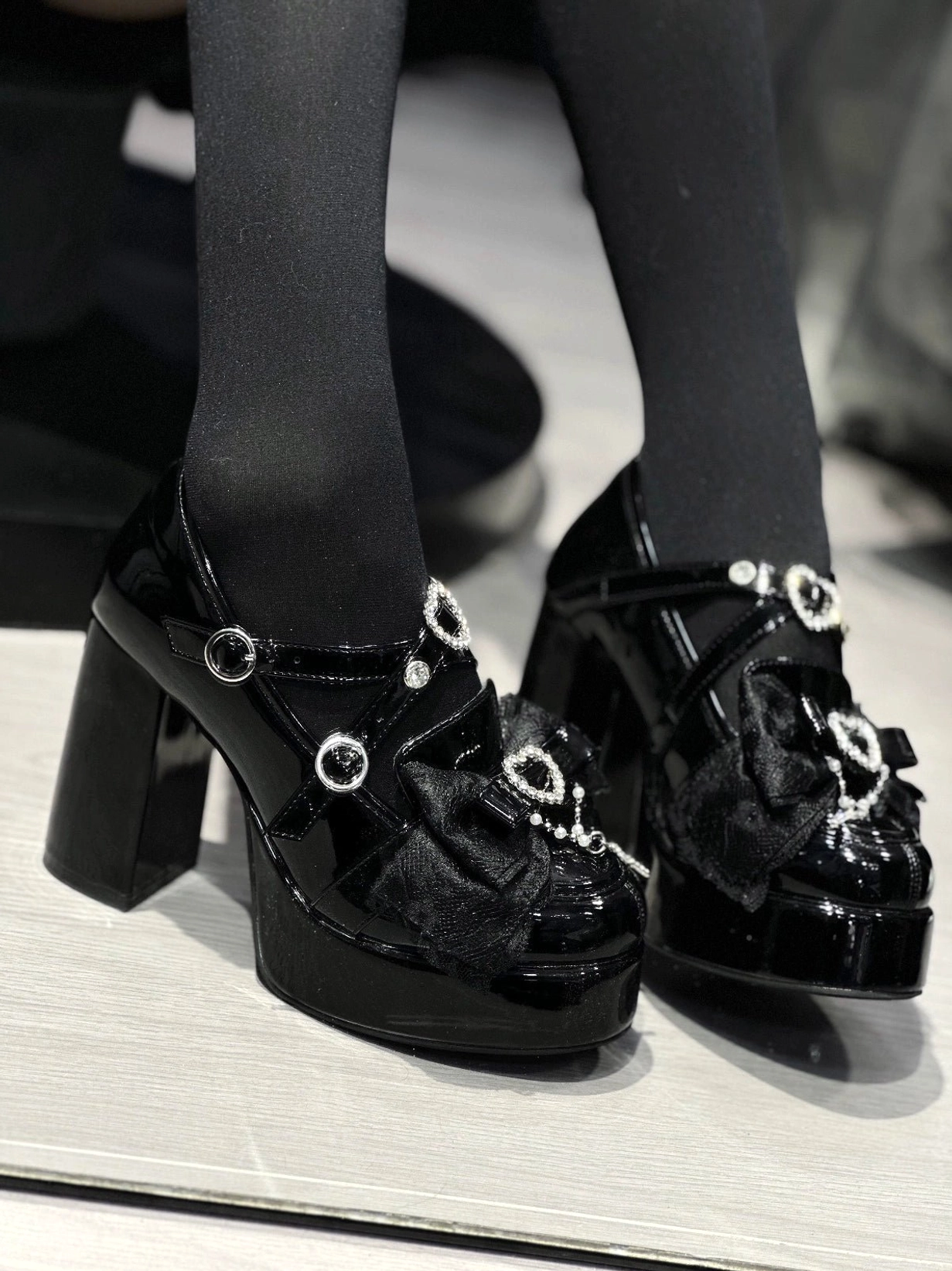 Side front close-up pair view of black Jirai Kei platform high-heel shoes with multiple heart rhinestone decorations, satin vamp bow, and thick platform heel structure
