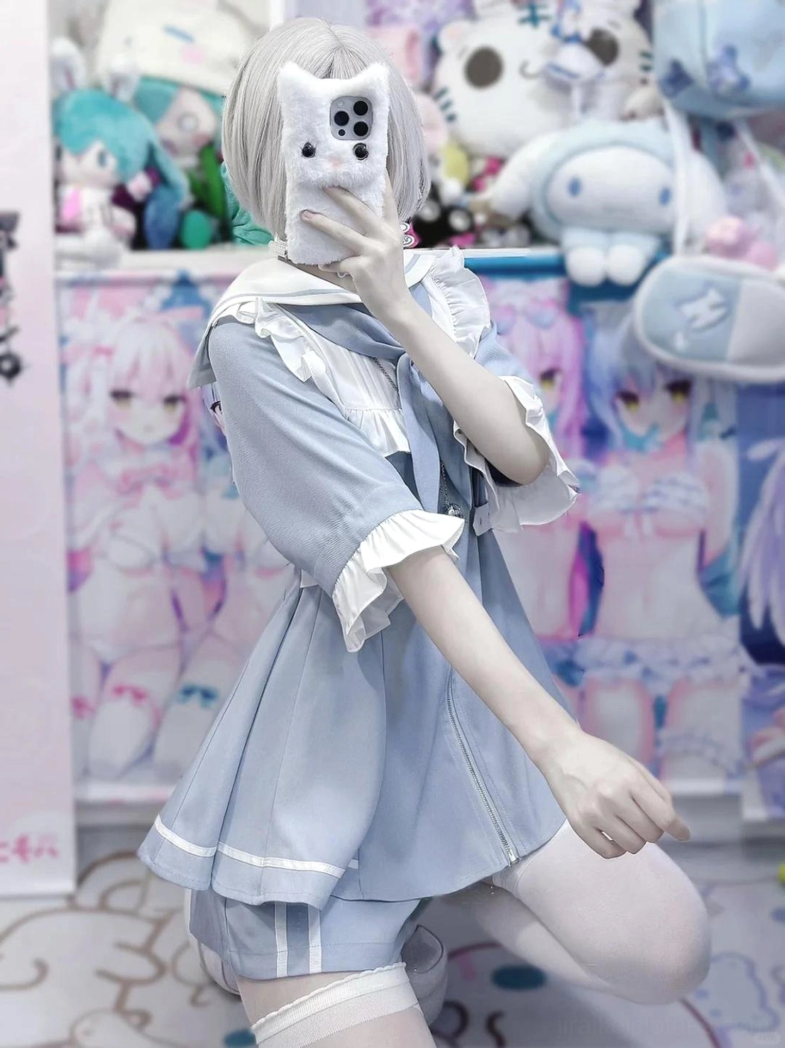 Low-angle selfie of pastel blue sailor Jirai Kei two-piece set featuring ruffled cuffs, front bow accent, matching shorts, and white thigh high sock against plush toy wall background