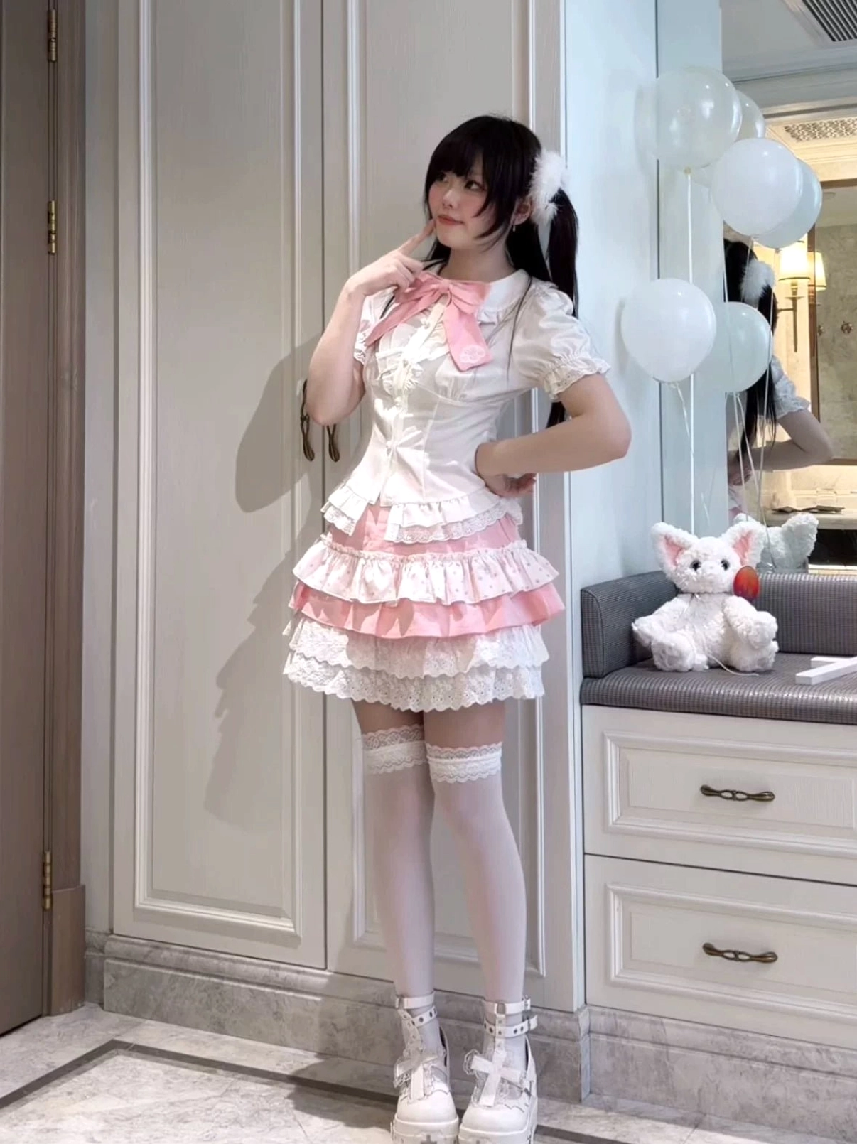 Full-body jirai kei sweet lolita dress outfit on model, short pink salopette dress with tiered ruffles, white thigh-high socks, pink bow tie collar, complete look