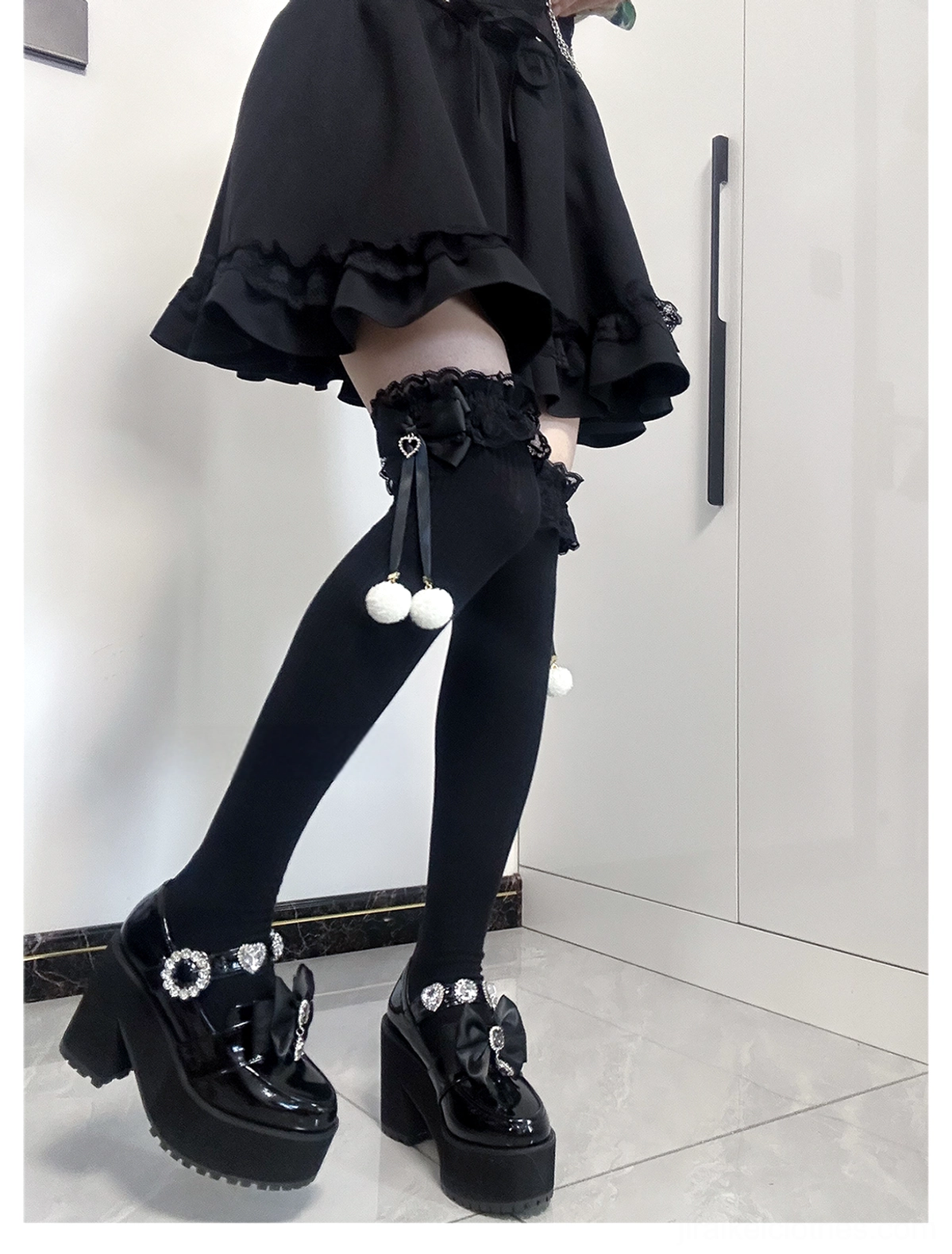Jirai Kei thigh-high socks three-quarter side posed view all-black solid socks lace cuffs black bow accents with white pom-pom charms indoor scene with platform shoes