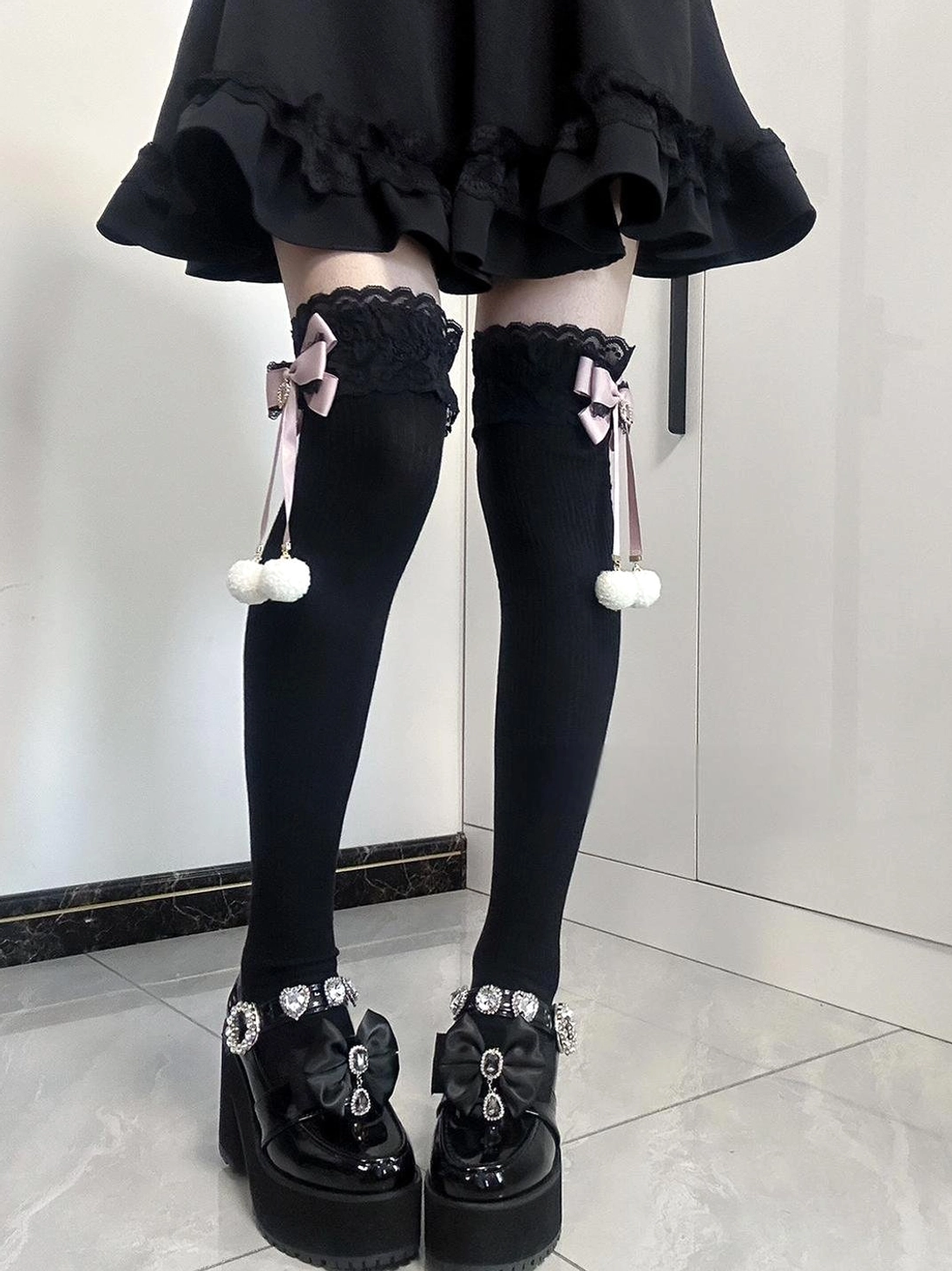 Matching Jirai Kei thigh-high socks symmetrical front leg view showing solid black socks with lace cuffs pink bows and cascading white pom-poms paired with black ruffled skirt and platform loafers