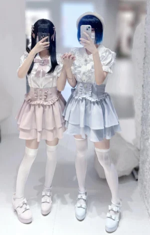 Two female models wearing jirai kei tiered mini skirts with matching blouses, full-body mirror selfie in boutique setting, showcasing ruffled details and bow accents