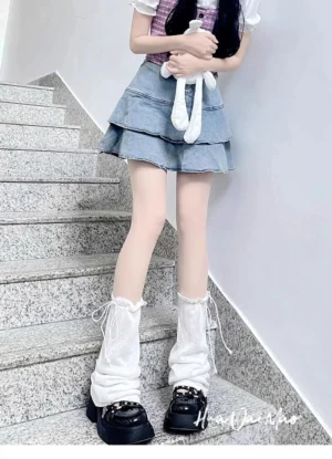 White Jirai Kei drawstring leg warmers with adjustable ankle ties worn by model in pink checkered top and layered denim skirt on indoor staircase, front full view