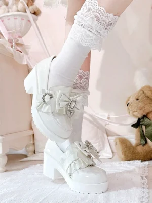 White patent leather Jirai Kei loafers side profile close-up showing chunky platform heel, pearl-like rhinestone bow decorations, and glossy high-shine finish with ruffled lace socks