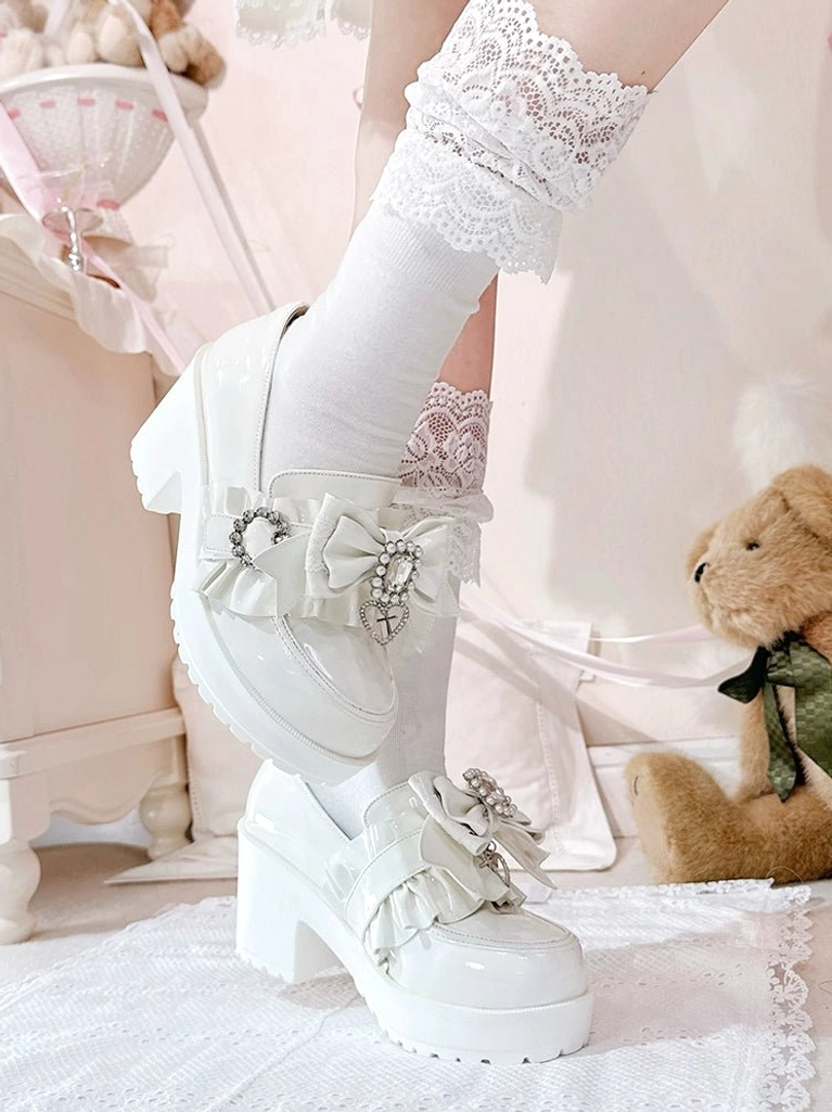 White patent leather Jirai Kei loafers side profile close-up showing chunky platform heel, pearl-like rhinestone bow decorations, and glossy high-shine finish with ruffled lace socks