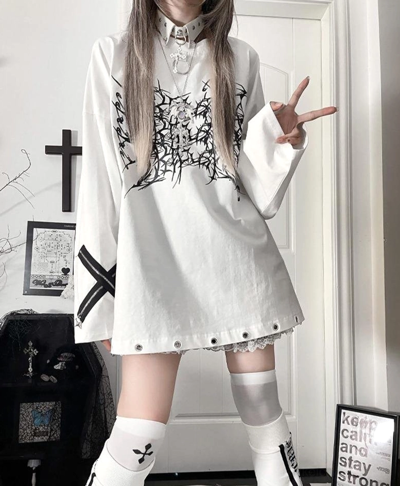 White oversized jirai kei gothic long-sleeve tee featuring black thorn cross print, cross strap sleeves, and riveted lace hem details, styled with thigh-high socks in alternative gothic room