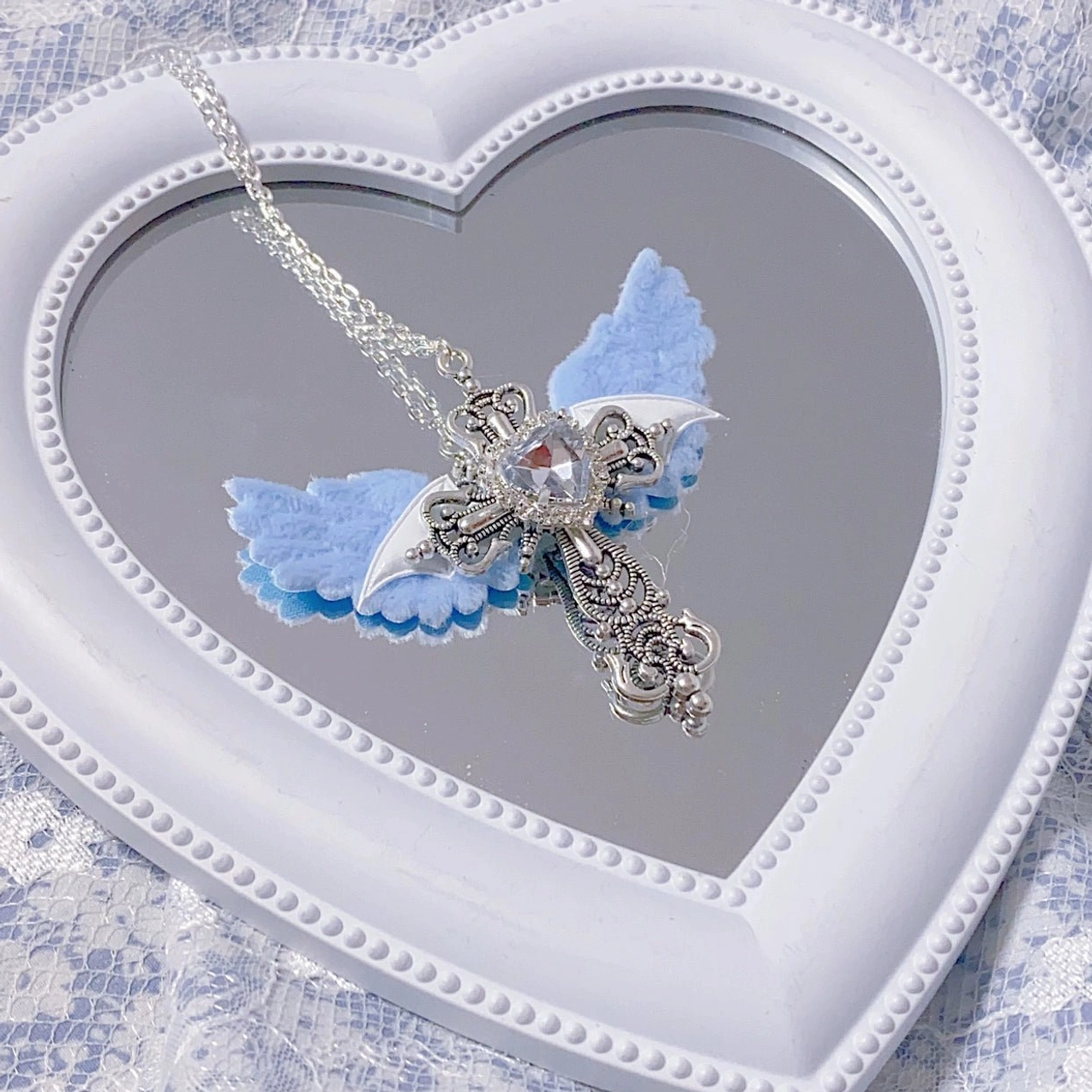 Light blue angel wing cross necklace with clear heart gem and silver ornate cross on white heart-shaped mirror with lace fabric backdrop