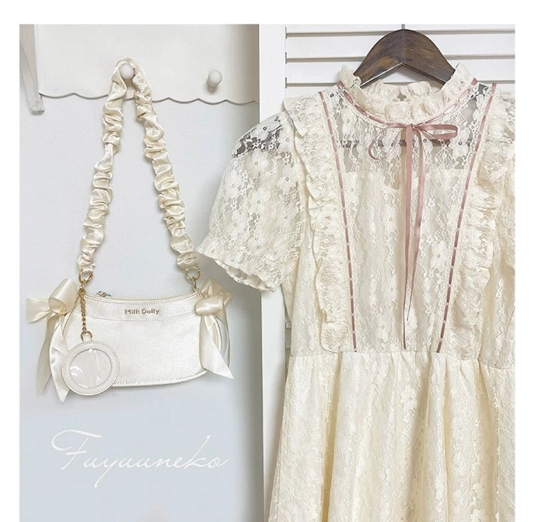 Mini detachable ita bag accessory and hanging display bag with ruched satin strap, bow charm, and round badge pendant shown in flat lay against lace dress background wall
