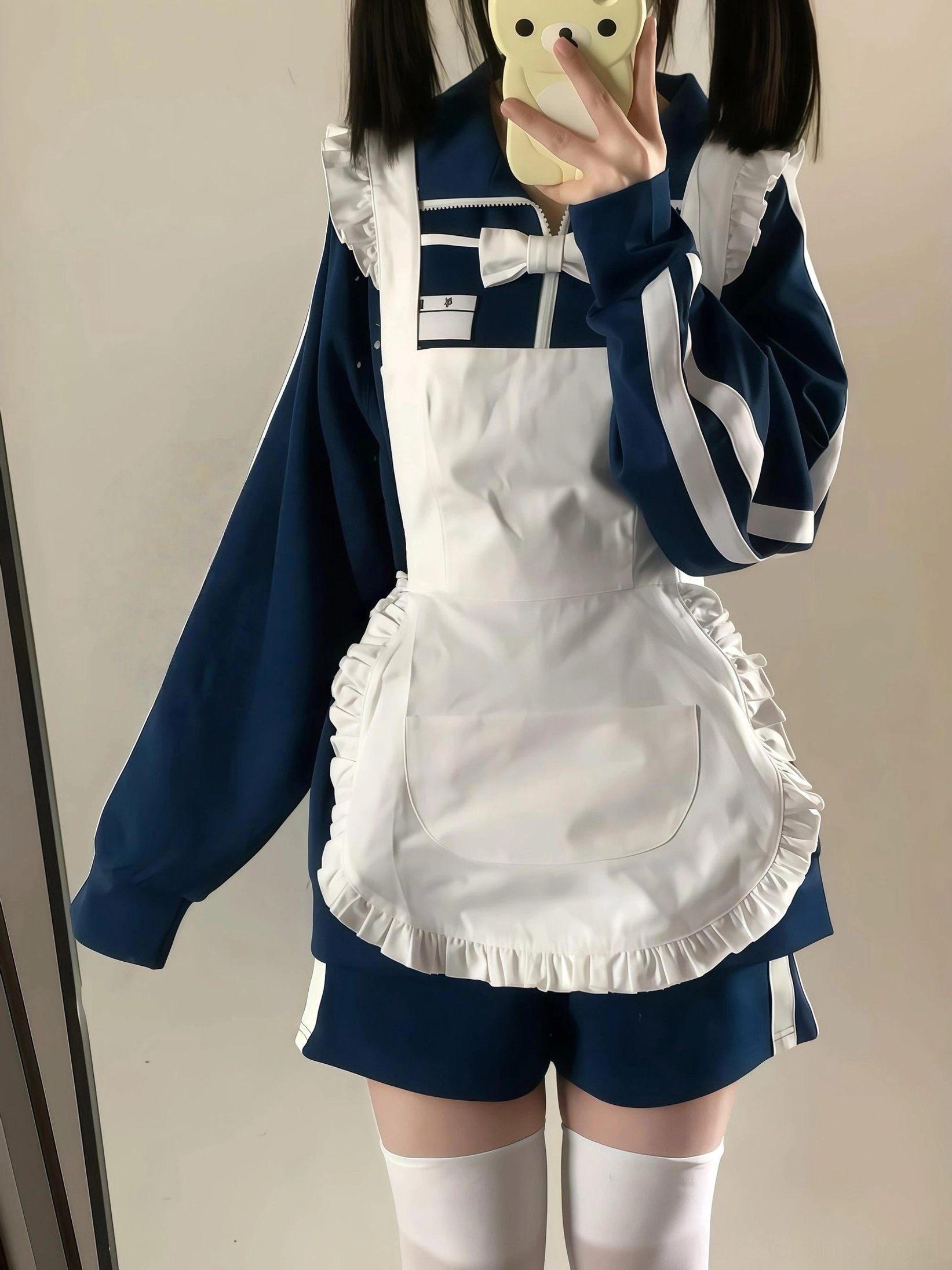 Navy Jirai Kei Jersey Maid Outfit worn by model with twin-tails hairstyle, featuring white ruffled maid apron with bow decoration and navy sports jacket with white sleeve stripes, indoor home setting